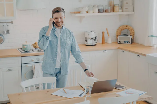 Bel homme barbu souriant parlant sur téléphone portable dans la cuisine tout en travaillant à distance — Photo