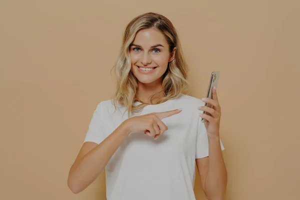 Cute excited female pointing on smartphone with forefinger and looking at camera with broad smile
