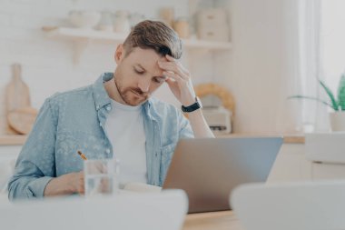 Mutfak masasında dizüstü bilgisayarla oturmuş notlar yazan odaklanmış bir adam.