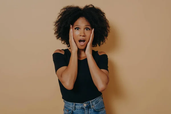 Photo de choquée jolie dame aux cheveux bouclés et à la peau foncée tenant la main sur les pommettes — Photo