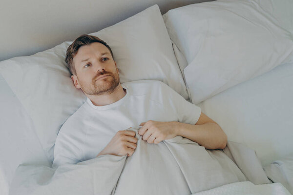 Lonely upset young bearded man lying in bed with open eyes