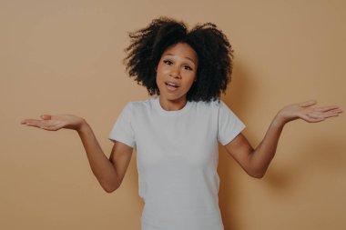 Emin olmayan Afrikalı kadın omuz silkiyor ve kollarını yana doğru açıyor, bej duvarın üzerinden izole edilmiş.