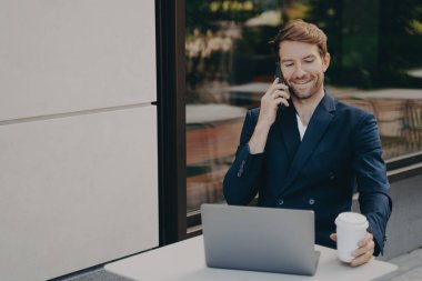 Genç başarılı işletme sahibi dışarıda kafede dizüstü bilgisayarda çalışıyor ve cep telefonuyla konuşuyor.