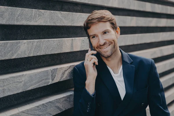 Bonito homem sorridente em elegante falando no smartphone fora, desfrutando de conversa — Fotografia de Stock