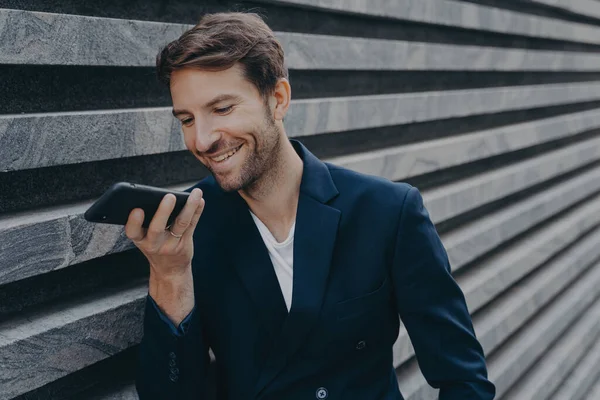 Jovem atraente homem fica do lado de fora e usando assistente de reconhecimento de voz no smartphone — Fotografia de Stock