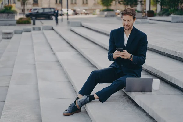 Jovem homem confiante em terno elegante usando smartphone enquanto sentado em passos fora com laptop — Fotografia de Stock