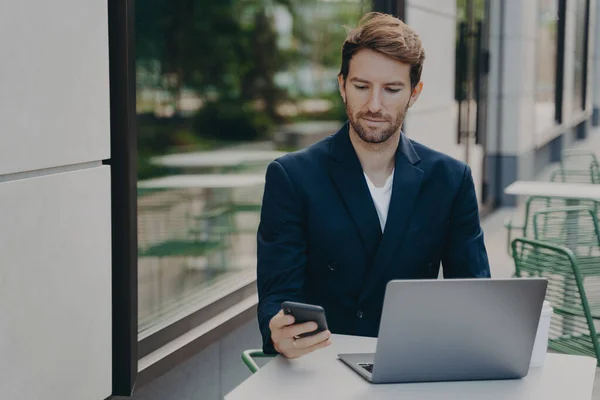 Jefe masculino serio vestido con ropa elegante funciona en línea en el ordenador portátil sostiene celular — Foto de Stock