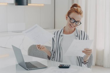 Evdeki masada oturan heyecanlı kızıl saçlı genç kadın, ev faturalarını kontrol ettikten sonra kendini mutlu hissediyor.,