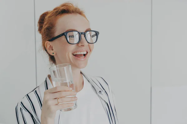 Feliz mulher de gengibre rindo de pé na cozinha de manhã com um copo de água mineral pura — Fotografia de Stock