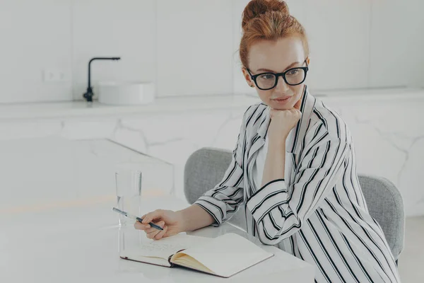 Ung koncentrerad ingefära kvinna tänker på en ny dag medan du gör anteckningar i anteckningsboken i köket — Stockfoto