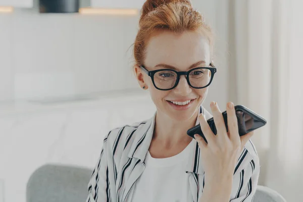 Vacker positiv rödhårig kvinna i glasögon som håller mobiltelefon och skicka ljudmeddelande — Stockfoto