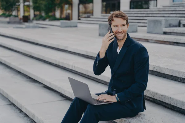 Joven empresario atractivo que trabaja en el ordenador portátil a distancia, hablando en el teléfono inteligente fuera — Foto de Stock