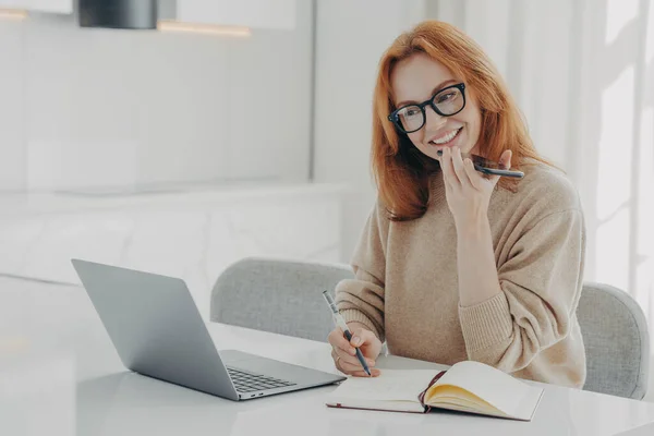 Mulher gengibre positiva em roupas casuais grava mensagem de áudio ou smartphone enquanto trabalhava no laptop — Fotografia de Stock