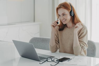 Bilgisayardaki video görüşmesi sırasında iş arkadaşlarıyla iletişim kuran, kulaklıklı, neşeli, kızıl saçlı bir kadın.