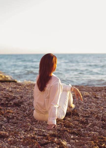 身穿白衬衫 面带微笑的年轻女子坐在海滨休息 观看海景落日 — 图库照片