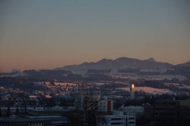 Arkasında dağ olan bir şehir. Gökyüzü turuncu ve güneş batıyor. Şehir ışıklarla aydınlanıyor.