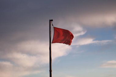 Red flag is flying in the sky. The sky is cloudy and the sun is setting. The flag is on a pole and is the only thing visible in the image