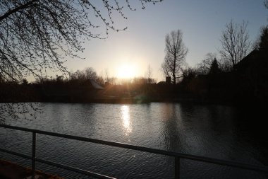 Yanı çitlerle çevrili sakin bir göl. Güneş batıyor ve su ışığı yansıtıyor.