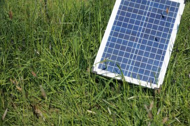 Çimlerin üzerinde bir güneş paneli var. Çimler yeşil ve gökyüzü mavi. Güneş paneli yenilenebilir enerjinin bir sembolüdür ve çimenler doğanın bir sembolüdür.