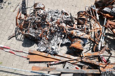 Bir araba yanıyor ve tahrip edildi. Sahne kaotik ve tehlikeli. Yangın çevreye yayıldı ve etrafa dağılmış metal ve enkaz parçaları var.