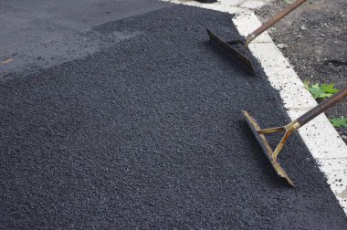 Two shovels are resting on a black surface, possibly a driveway or a road. The shovels are made of metal and have a black handle. The surface is covered in a layer of black gravel or asphalt