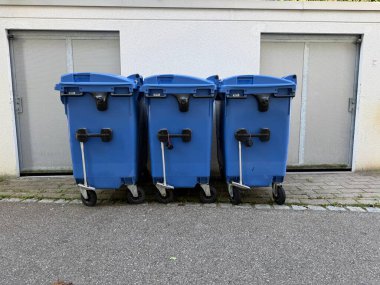 Üç mavi çöp tenekesi duvara karşı dizilmiş. Çöp tenekeleri mavi ve tekerlekleri var.