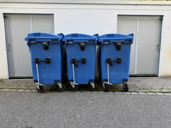 Üç mavi çöp tenekesi duvara karşı dizilmiş. Çöp tenekeleri mavi ve tekerlekleri var.