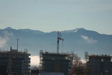 İskeleli birden fazla bina ve önemli bir vinç bulunan bir inşaat alanının geniş bir görüntüsü, uzak, karlı dağların ve puslu bir vadinin zeminine kurulmuş..
