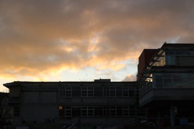 Çarpıcı geniş bir modern bina görüntüsü dramatik bir günbatımı gökyüzü karşısında canlı turuncu ve sarı bulutlar karamsar ve atmosferik bir manzara yaratıyor..