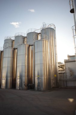 Sıralar dolusu parlak, paslanmaz çelik sanayi siloları ve güvenlik merdivenleri parlak mavi gökyüzüne karşı güneş ışığını yansıtıyor. Çeşitli sıvılar ya da malzemeler için modern depolama tankları.