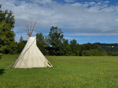 Çadır arka planda ağaçlar olan çimenli bir tarlada. Çadır beyazdır ve koni şeklindedir. Tarla yemyeşil, gökyüzü açık ve mavi.