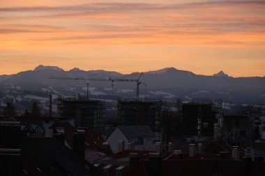 İnşaat halindeki binaların üzerinde günbatımının ve arka planda dağların olduğu panoramik bir manzara.