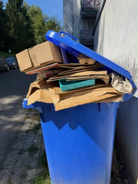 Mavi çöp kutusu çöp dolu, karton kutular ve kağıt da dahil. Çöp tenekesi taşmış ve kapağı da kapalıymış. Olay yeri, bölgenin uygun bir atık yönetimine ihtiyacı olduğunu gösteriyor.
