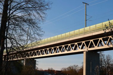 Yeşil korkuluklu ve arka planda ağaç olan bir köprü. Köprü bir yolun üzerinde ve gökyüzü açık.