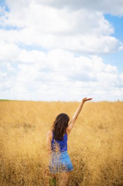 Mavi tişörtlü ve kot şortlu güzel esmer bir kadının portresi tarlanın ortasında duruyor, açık havada bir yaz gününün arka planında yüzünü güneşe gösteriyor.