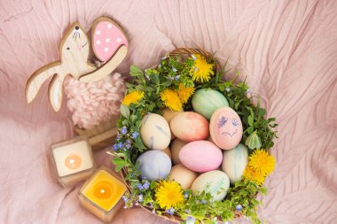 Pastel bir yatak örtüsü üzerinde bir sepet çimen ve çeşitli parlak çiçekler var, bir tavşan. Sepette suluboya güzel pastel renklerle boyanmış Paskalya yumurtaları var.