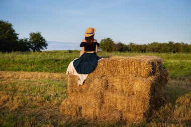 Hasır şapkalı genç bir kadın batan güneşe bakıyor, kız büyük bir saman yığını üzerinde oturuyor..