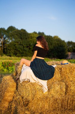 Hasır şapkalı genç bir kadın batan güneşe bakar, kız büyük bir saman yığınının üzerinde oturur. Sıcak yaz akşamı