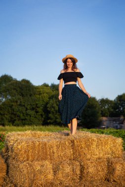 Hasır şapkalı güzel bir kadın büyük bir saman balyasının üzerinde duruyor. Özgürlük, rüzgar, batan güneş