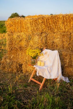 Tarlada saman balyalarının yanında piknik. Batan güneş. Kırsal tarzda - ahşap sandalye, ekose, çiçek buketi, mumlar