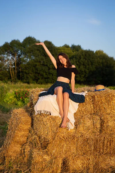 Genç ve güzel bir kadın batan güneşe bakar ve elini sallar, büyük bir saman yığınının üzerinde oturur.
