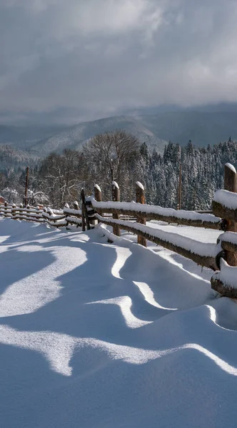 风景如画的影子在雪地上从木栅栏上飘扬 高山冬季小村子郊外 雪地小径 长满了冷杉 长满了薄雾 多云的小山 — 图库照片