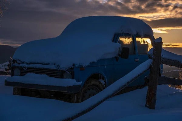 Snow covered car on small and quiet mountain village outskirts. Dusk winter sunrise, Carpathian, Ukraine. Car model old and unrecognizable.