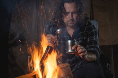 Ateşin yanında kamp yapan bir adam konserve yiyecekler yiyor..