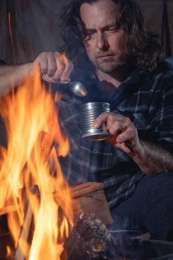 Ateşin yanında kamp yapan bir adam konserve yiyecekler yiyor..