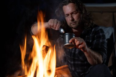 Ateşin yanında kamp yapan bir adam konserve yiyecekler yiyor..