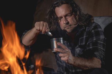 Ateşin yanında kamp yapan bir adam konserve yiyecekler yiyor..