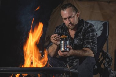 Ateşin yanında kamp yapan bir adam konserve yiyecekler yiyor..