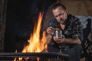 Ateşin yanında kamp yapan bir adam konserve yiyecekler yiyor..