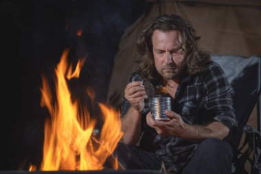 Ateşin yanında kamp yapan bir adam konserve yiyecekler yiyor..